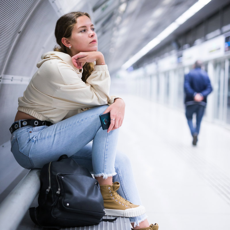 Young woman waiting for a train with a backpack at her feet and a phone in her hand.