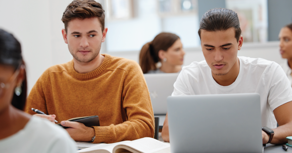 Two students sitting side-by-side at a table, with one student looking out of the corner of his eye at the other student's laptop computer.