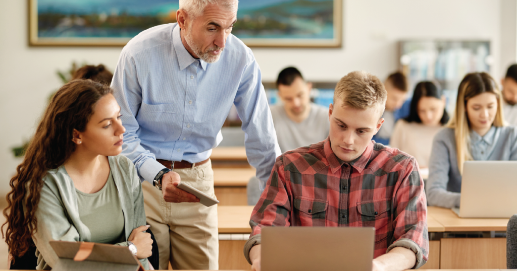 Classroom setting, with students seated at long tables and working on laptop computers. In the foreground, an instructor is helping a student while another student looks on.